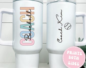 May include: White insulated tumbler with a handle. One side features the word "COACH Baseball" in pastel colors. The other side has the name "Coach Kim" with a baseball graphic. The tumbler has a clear lid and a pink sticker that says "Prints Both Sides".