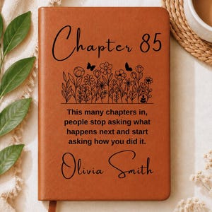 May include: A tan leather-bound journal featuring the black text 'Chapter 85' above a line drawing of wildflowers and butterflies. Below the flowers, the text reads 'This many chapters in, people stop asking what happens next and start asking how you did it.' followed by the name 'Olivia Smith'.