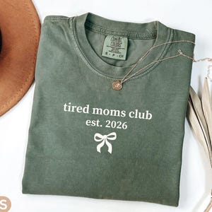 May include: A moss green t-shirt with the text "tired moms club est. 2026" and a bow graphic. A gold-colored necklace with a pendant and a brown hat are also visible. The shirt is made of a soft material.
