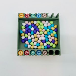 Op de afbeelding: Een groene houten spelorganizer gevuld met kleurrijke, druppelvormige spelstukken. De organizer heeft sleuven voor fiches met diverse voedselgerelateerde iconen. De stukken zijn in tinten blauw, roze, geel en groen.