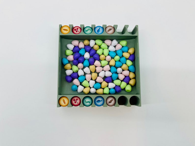 Op de afbeelding: Een groene houten spelorganizer gevuld met kleurrijke, druppelvormige spelstukken. De organizer heeft sleuven voor fiches met diverse voedselgerelateerde iconen. De stukken zijn in tinten blauw, roze, geel en groen.