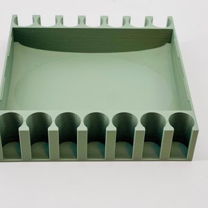 Op de afbeelding: Een saliegroene, rechthoekige organizer met een reeks cilindrische sleuven langs &eacute;&eacute;n kant en een groter, open compartiment. De bovenrand heeft een reeks kleine, rechthoekige uitsteeksels. Het item is gemaakt van een effen, matte materiaal.