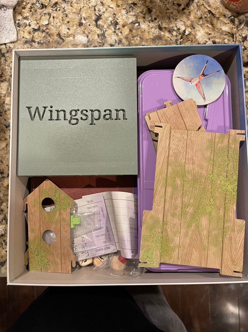 Op de afbeelding: Open doos van het bordspel Wingspan, met een groene doos met de titel, houten spelstukken, een paarse container en een rond spelstuk met een vogelillustratie.