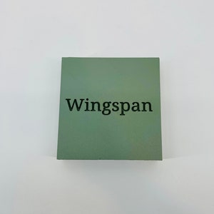 Op de afbeelding: Een vierkant, saliegroen object met het woord "Wingspan" in zwart gedrukt. Het object is gecentreerd op een witte achtergrond.