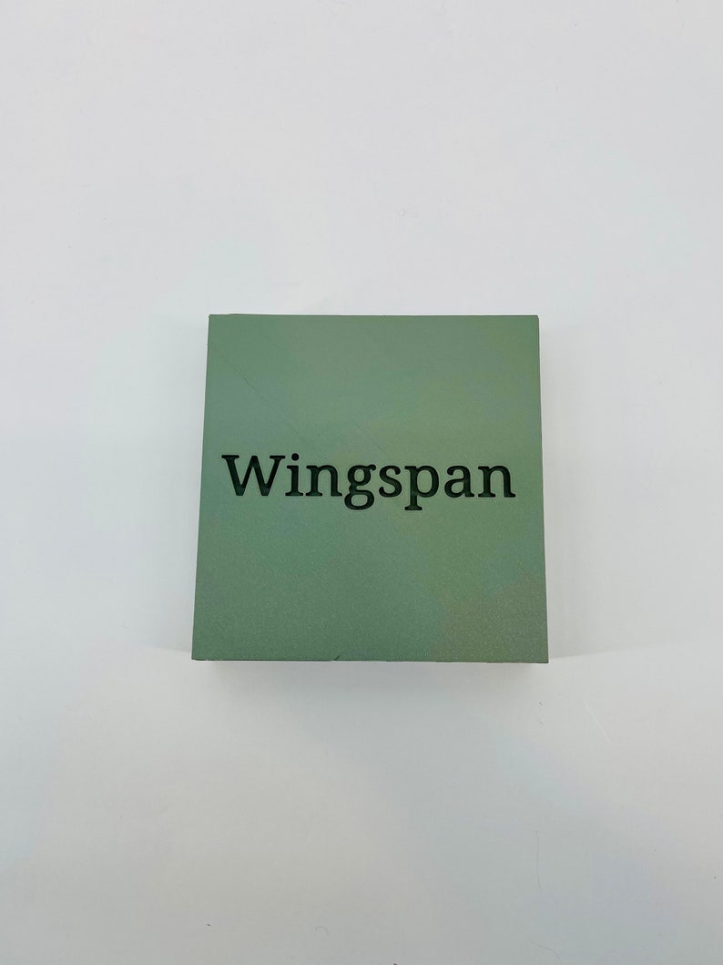 Op de afbeelding: Een vierkant, saliegroen object met het woord "Wingspan" in zwart gedrukt. Het object is gecentreerd op een witte achtergrond.
