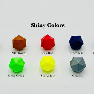 Op de afbeelding: Een verzameling van tien veelkleurige dobbelstenen in glanzende kleuren, waaronder Glitter Red, Silk Bronze, Silk Red, Glitter Blue, Silk Blue/Purple, Glitter Green, Grass Green, Silk Yellow, Chrome en Yellow Gold. De tekst "Shiny Colors" is zichtbaar.
