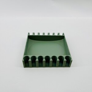 Op de afbeelding: Een saliegroene organizer met meerdere cilindrische sleuven en een rechthoekige basis. De bovenrand heeft gelijkmatig verdeelde verticale verdelers. Het product is ontworpen om kleine items zoals pennen of gereedschap te bevatten.