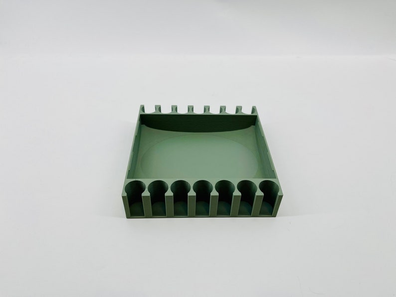 Op de afbeelding: Een saliegroene organizer met meerdere cilindrische sleuven en een rechthoekige basis. De bovenrand heeft gelijkmatig verdeelde verticale verdelers. Het product is ontworpen om kleine items zoals pennen of gereedschap te bevatten.