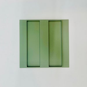 Op de afbeelding: Een vierkant, saliegroen decoratief dienblad met twee verticale rechthoekige uitsparingen. De lade is gemaakt van een massief materiaal en staat op een witte achtergrond. Het ontwerp is minimalistisch en modern, geschikt voor woondecoratie.