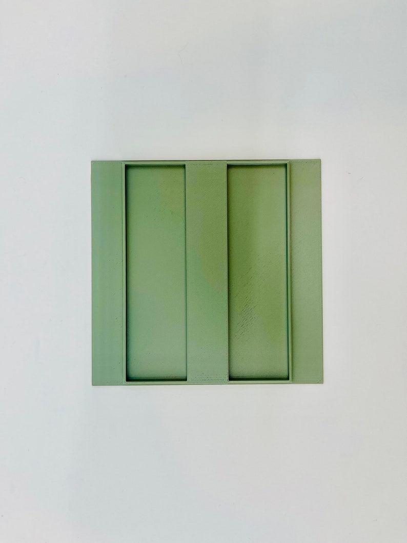 Op de afbeelding: Een vierkant, saliegroen decoratief dienblad met twee verticale rechthoekige uitsparingen. De lade is gemaakt van een massief materiaal en staat op een witte achtergrond. Het ontwerp is minimalistisch en modern, geschikt voor woondecoratie.