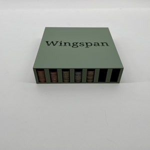 Op de afbeelding: Een saliegroene houten spelorganizer voor het bordspel Wingspan. De organizer heeft sleuven gevuld met stapels speltoken. Het woord "Wingspan" staat erop gedrukt.
