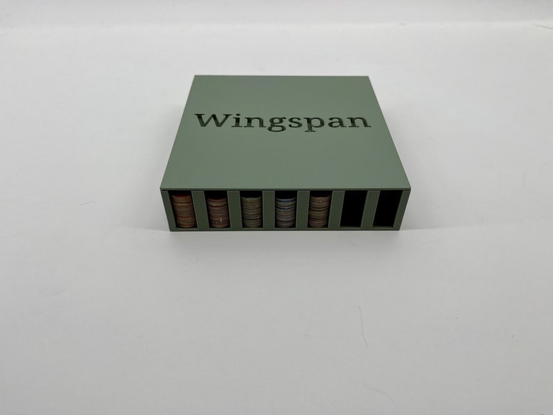 Op de afbeelding: Een saliegroene houten spelorganizer voor het bordspel Wingspan. De organizer heeft sleuven gevuld met stapels speltoken. Het woord "Wingspan" staat erop gedrukt.