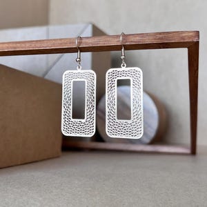 May include: A pair of silver rectangle-shaped earrings with a delicate, intricate design. The earrings have a geometric pattern and are hanging from a silver hook.