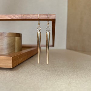 以下が含まれることがあります： 先が尖った涙滴型のデザインの金色のイヤリングのペア。イヤリングは木製の表面にぶら下がっています。