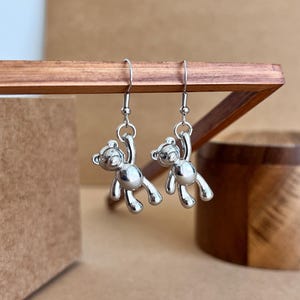 Op de afbeelding: Zilveren oorhangers met een klein berenbedeltje. De beer hangt aan de oorhaak en heeft een glanzende afwerking.