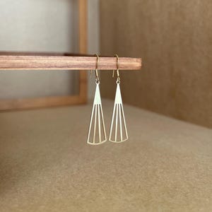 May include: A pair of gold-toned earrings with a geometric, fan-shaped design. The earrings are hanging from a wooden surface.