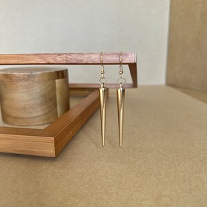 以下が含まれることがあります： 長い先のとがったデザインの金色のイヤリングのペア。イヤリングは木製の表面にぶら下がっています。