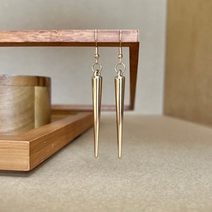 May include: A pair of gold-toned earrings with a long, pointed design. The earrings are hanging from a wooden surface.