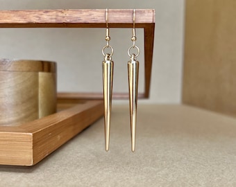 May include: A pair of gold-toned earrings with a long, pointed design. The earrings are hanging from a wooden surface.