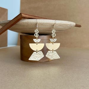 May include: Gold geometric earrings with a hammered texture. The earrings feature a crescent moon shape, a diamond shape, and a swirl design.