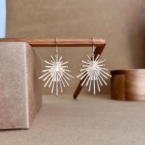 Puede incluir: Un par de pendientes dorados en forma de estrella. Cada pendiente presenta un diseño circular central con líneas radiales, creando un efecto de sol. Los pendientes están suspendidos de ganchos simples, mostrando un diseño moderno.