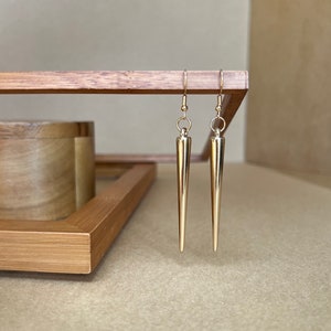 以下が含まれることがあります： 先のとがった涙型の金色のイヤリングのペア。イヤリングは木製の表面にぶら下がっています。