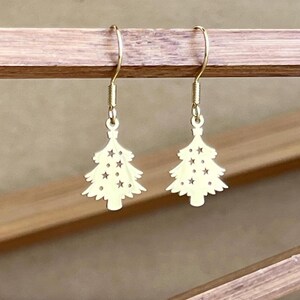 May include: A pair of gold-colored Christmas tree earrings. Each earring features a cutout tree design with star-shaped cutouts. The earrings hang from gold-colored ear wires.