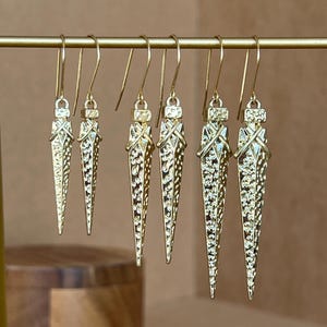 Pode incluir: Cinco pares de brincos dourados com um design pontiagudo texturizado. Os brincos são exibidos em um suporte dourado.