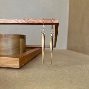 以下が含まれることがあります： 長い先のとがったデザインの金色のイヤリングのペア。イヤリングは木製の表面からぶら下がっています。