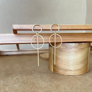 Puede incluir: Un par de pendientes de color dorado con una barra larga y fina y dos círculos. Los círculos están conectados a la barra y están ligeramente desplazados uno del otro.