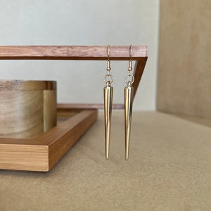 以下が含まれることがあります： 先が尖った細長いデザインの金色のイヤリングのペア。イヤリングは木製の表面にぶら下がっています。