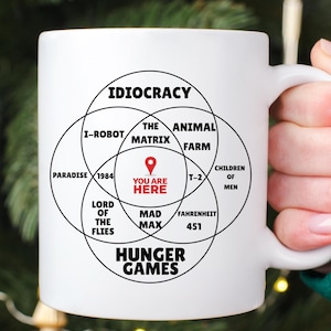 Può includere: Tazza in ceramica bianca con un diagramma di Venn. I cerchi sono etichettati con titoli di fantascienza come "Idiocracy" e "Hunger Games". Il testo "You Are Here" è al centro.