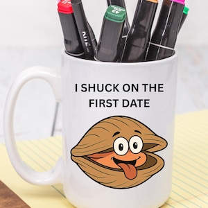 Puede incluir: Taza de cerámica blanca con el texto "I Shuck on the First Date" y una ilustración de dibujos animados de una almeja. La taza contiene varios rotuladores y lápices negros. La taza está sobre un bloc de notas amarillo.