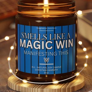 May include: A brown glass candle jar with a blue label that reads "Smells Like A Magic Win Manifesting This W All-Natural Soy Candle Christmas Warmth 9 oz". The candle is lit and the flame is visible.