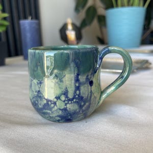 Op de afbeelding: Een handgemaakte keramische mok met een afgeronde basis en een gebogen handvat. De mok heeft een gemarmerd ontwerp in tinten van blauwgroen, groen en marineblauw. De binnenkant is effen blauwgroen. De mok staat op een wit oppervlak.