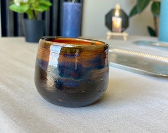 Peut inclure: Un gobelet en céramique fait à la main avec une finition brillante et multicolore. Les couleurs passent d'une base sombre, presque noire, à des nuances de bleu, de marron et de rouge-orangé sur le bord. La petite tasse arrondie est posée sur une surface blanche.