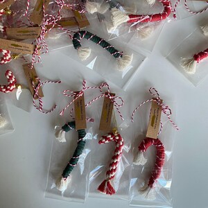 May include: A collection of handmade candy cane decorations, each wrapped in a clear plastic bag with a red and white twine bow. The decorations are made of red and white rope, green and white rope, or red and white rope with a white pom-pom. Each decoration has a small tag attached with the text "Happy Holidays Love, Jackie".