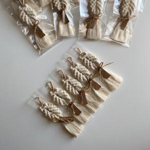 Pode incluir: Um grupo de chaveiros de macramé branco com argolas douradas e borlas. Os chaveiros são embalados individualmente em sacos de plástico transparentes com uma pequena etiqueta castanha que diz "Knotty Nice".