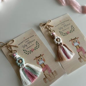 Puede incluir: Dos favores de baby shower con un diseño de jirafa. Cada favor incluye un pequeño llavero de borla hecho a mano con un dije de flor. La tarjeta dice "Bienvenido a la fiesta de bienvenida al bebé en honor a Lily-Maria el 11 de septiembre de 2023".