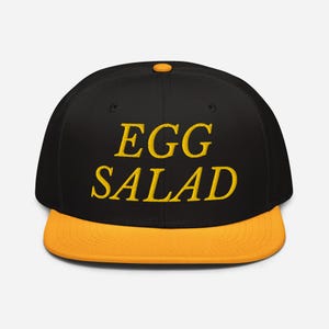 Può includere: Cappellino da baseball nero e giallo con la scritta ricamata "EGG SALAD". Il cappellino ha una visiera piatta e un bottone sulla parte superiore. Il testo è in grassetto giallo.