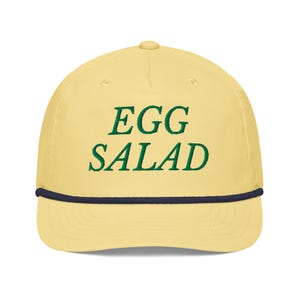Könnte beinhalten: Hellgelbe Baseballkappe mit dem gestickten Schriftzug "EGG SALAD" in Grün. Die Kappe hat eine marineblaue Kordel am Rand.