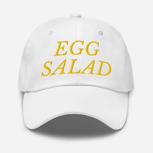 Op de afbeelding: Witte baseballpet met de tekst "EGG SALAD" geborduurd in geel.