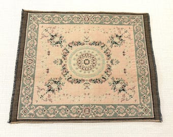 May include: A square woven textile mat featuring a pale pink center with a central circular floral medallion in sage green and cream. The border displays a repeating scroll pattern in sage green, framed by a dark brown edge with frayed fringe.