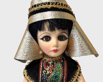 May include: A vintage doll with black hair and large eyes, wearing a gold-colored headdress and a black dress with gold trim. The dress features an embroidered paisley design in red, green, and gold. The doll has a painted face with red lips.