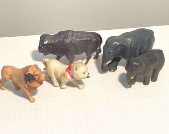May include: Five plastic animal figurines: a brown lion, a white dog with a red ribbon, a black buffalo, a gray elephant, and a smaller gray elephant.
