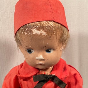 May include: A vintage porcelain doll with blonde hair wearing a red cap and a red shirt with a black bow tie.