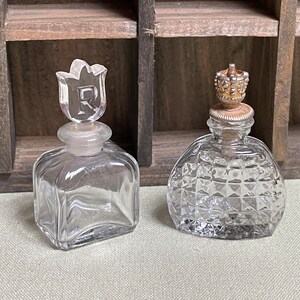 May include: Two clear glass perfume bottles with decorative stoppers. The bottle on the left has a clear glass stopper shaped like a tulip with the letter "R" etched into it. The bottle on the right has a gold-colored metal stopper with a crown design.