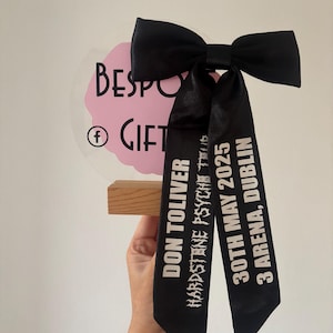 Könnte beinhalten: Ein schwarzes Satinband mit weißem Text "Don Toliver", "Hardstone Psycht", "30th May 2025" und "3 Arena, Dublin", das zu einer Schleife gebunden ist, die um einen durchsichtigen Acrylkreisel mit einer rosa Wolke und dem Text "Bespo Gift" und einem Facebook-Symbol gebunden ist.