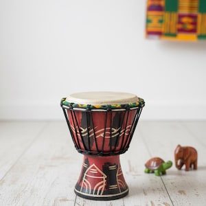 Könnte beinhalten: Eine kleine, handbemalte Djembe-Trommel mit roter Basis und schwarzen Akzenten. Das Trommelfell ist hellbraun und der Rand ist mit grünem, gelbem und schwarzem Seil verziert. Eine Holzschildkröte und ein Elefant sind im Hintergrund.