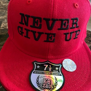 Puede incluir: Gorra de b&eacute;isbol roja con un mensaje bordado en negro que dice "NEVER GIVE UP". La gorra tiene una etiqueta negra y plateada con el texto "KB ETHOS PREMIUM HEADWEAR" y "7 1/8 SIZE".